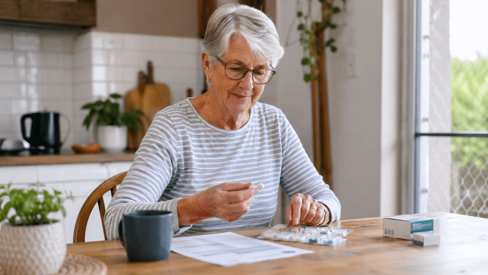 An older woman at home at her kitchen table reading a prescription label with medicines laid out beside her.
