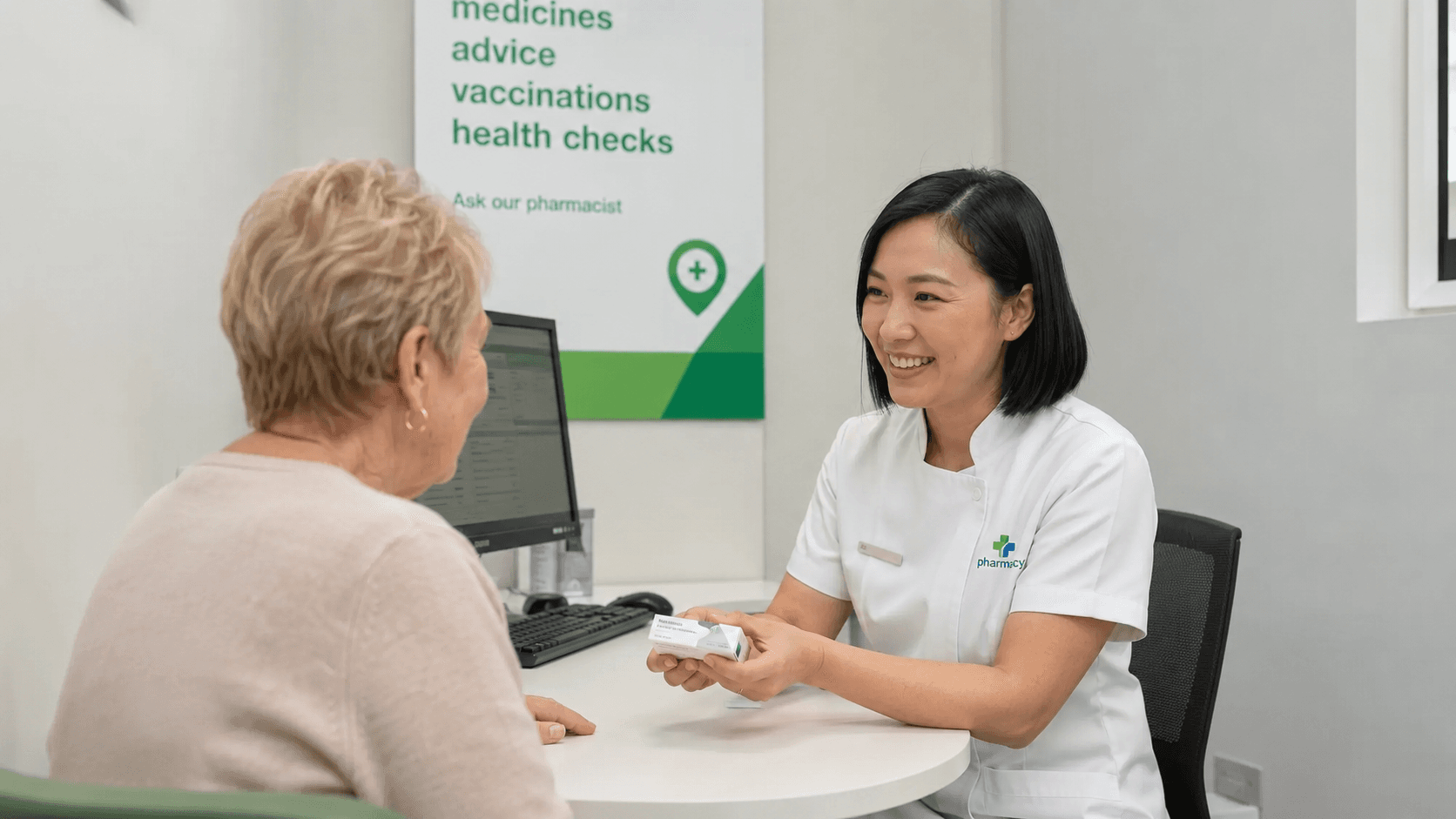 A pharmacist in a private consultation room with an older female patient, beside a sign listing medicines, vaccinations and health checks.
