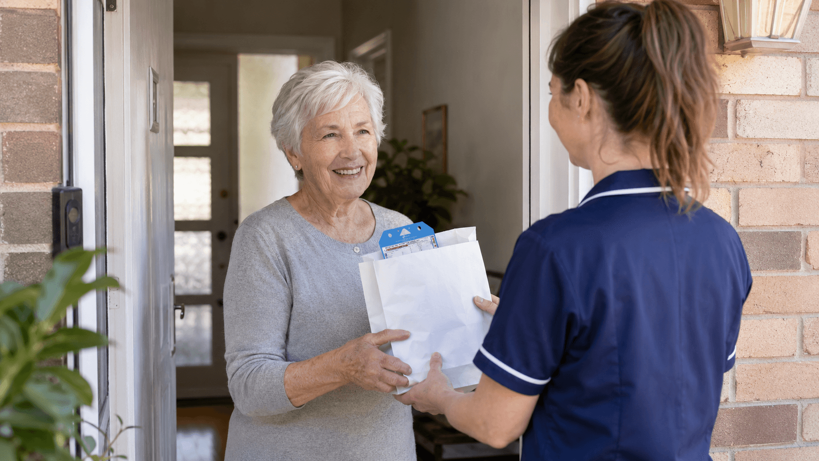 A pharmacy nurse handing medicines and paperwork to an older woman at the front door of her home in Australia.