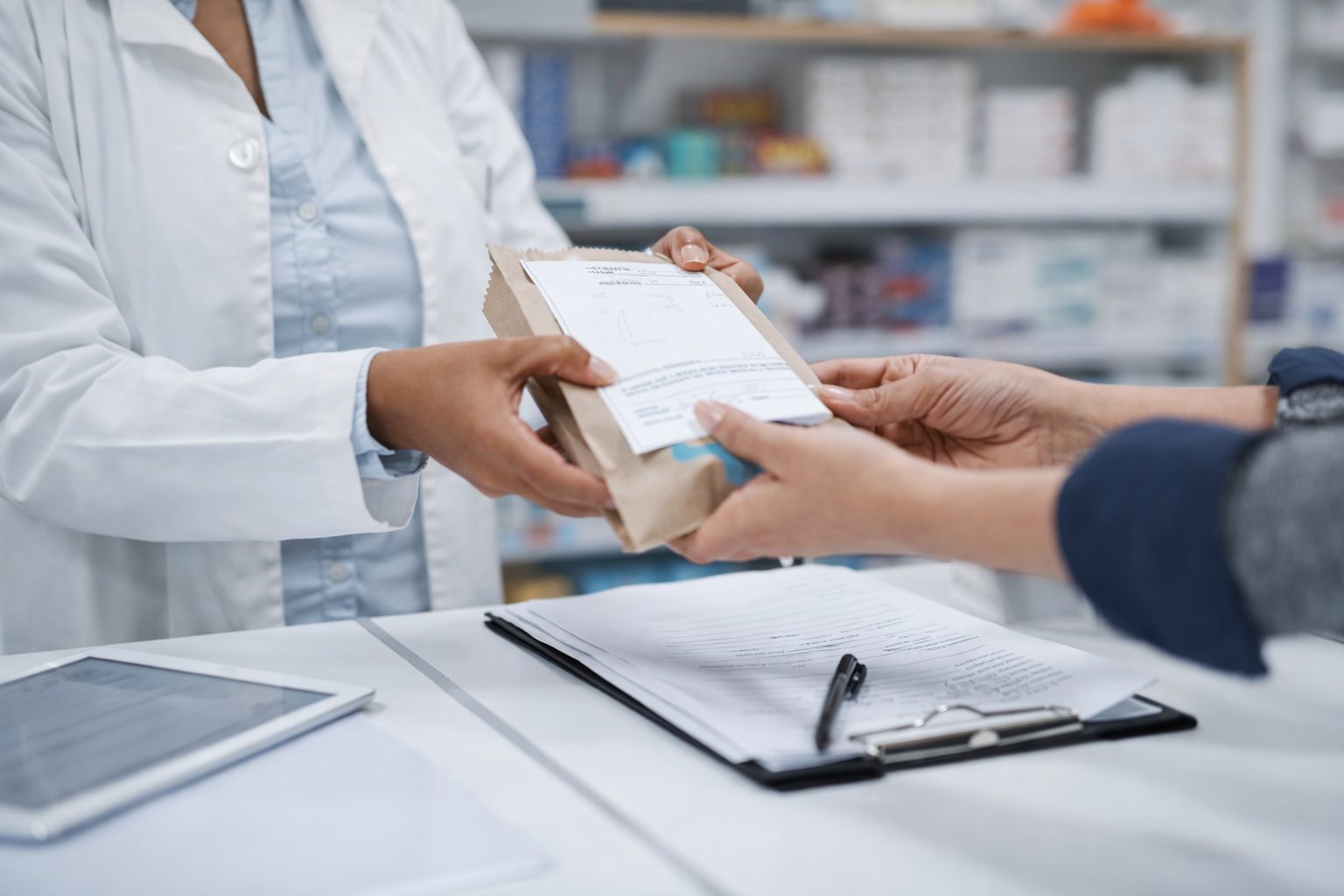A pharmacist handing a paper prescription bag to a customer across the counter of an Australian pharmacy.